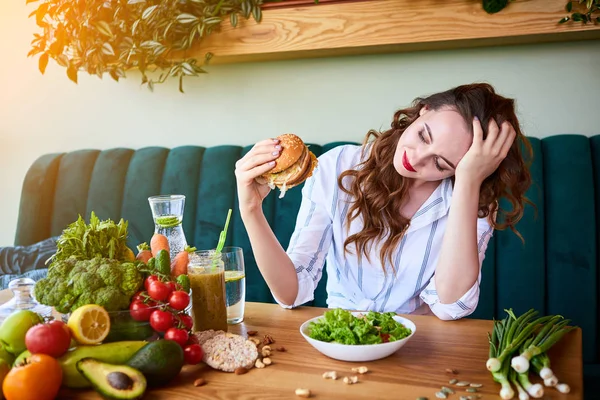 Όμορφη νεαρή γυναίκα αποφασίζει να φάει χάμπουργκερ ή φρέσκια σαλάτα στην κουζίνα. Φτηνές πρόχειρο φαγητό εναντίον υγιεινής διατροφής — Φωτογραφία Αρχείου