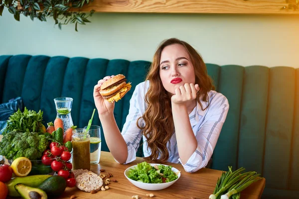 美しい若い女性はキッチンでハンバーガーや新鮮なサラダを食べることを決定します.安いジャンクフード対健康的な食事 — ストック写真