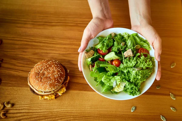 Γυναίκα αποφασίζει να φάει χάμπουργκερ ή φρέσκια σαλάτα στην κουζίνα. Φτηνές πρόχειρο φαγητό εναντίον υγιεινής διατροφής — Φωτογραφία Αρχείου