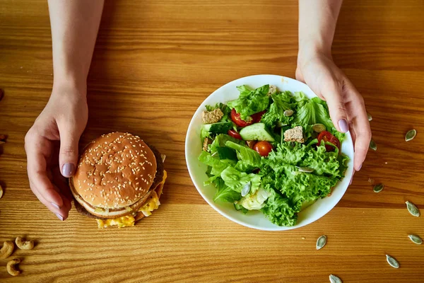 Γυναίκα αποφασίζει να φάει χάμπουργκερ ή φρέσκια σαλάτα στην κουζίνα. Φτηνές πρόχειρο φαγητό εναντίον υγιεινής διατροφής — Φωτογραφία Αρχείου