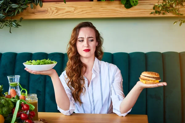 美しい若い女性はキッチンでハンバーガーや新鮮なサラダを食べることを決定します.安いジャンクフード対健康的な食事 — ストック写真