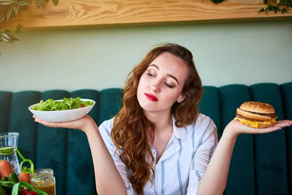 Красивая молодая женщина решает есть гамбургер или свежий салат на кухне. Дешевая вредная пища против здорового питания — стоковое фото