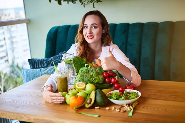 Woman dietitian in medical uniform with tape measure working on a diet plan sitting with different healthy food ingredients in the green office on background. Weight loss and right nutrition concept