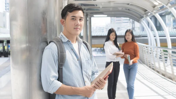 Energetic optimistic teen man with stationary and book stand front the student girl people.