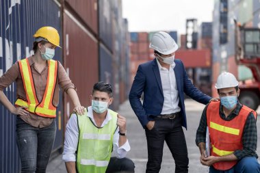 Grup çalışanları koruyucu maske ve güvenlik kaskı takıyor ve konteyner kargo deposunun arka planına sahip takım elbise giyiyor. Çalışan endüstri çalışanı kavramı.