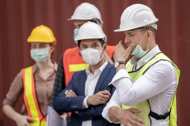 İş adamı ve fabrika işçileri tıbbi maske takıyor ve açık hava fabrikası kargo deposunda güvenlik elbisesi giyiyor..