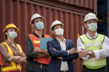 İş adamı ve fabrika işçileri tıbbi maske takıyor ve açık hava fabrikası kargo deposunda güvenlik elbisesi giyiyor..