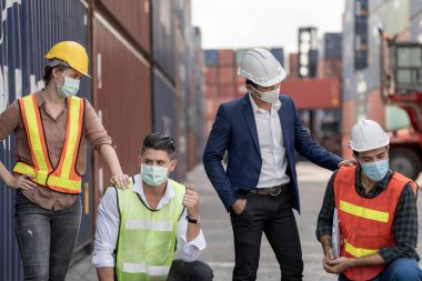 Grup çalışanları koruyucu maske ve güvenlik kaskı takıyor ve konteyner kargo deposunun arka planına sahip takım elbise giyiyor. Çalışan endüstri çalışanı kavramı.