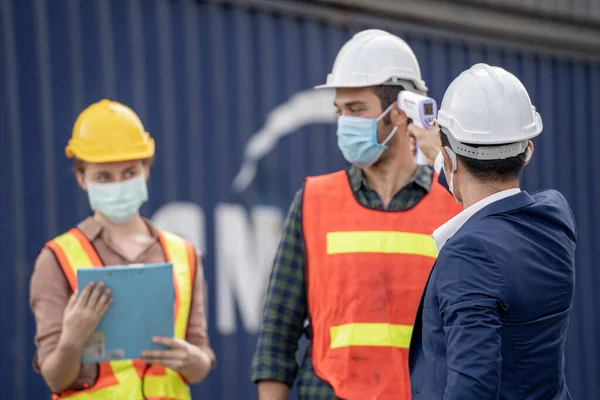 Factory man worker in a medical mask and safety cloth use equipment measures the worker temperature with a non-contact infrared thermometer