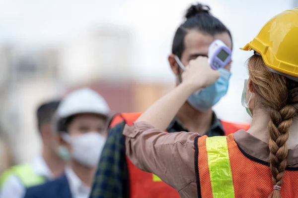 Factory woman worker in a face medical mask and safety dress used measures temperature at worker people standing on queue with a non-contact infrared thermometer