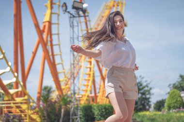 Lunaparktaki lunaparkta hız treninin önünde mutlu bir şekilde duran genç turist kadın. Yaz günü tatilde mutlu bir hayatı kabul et..