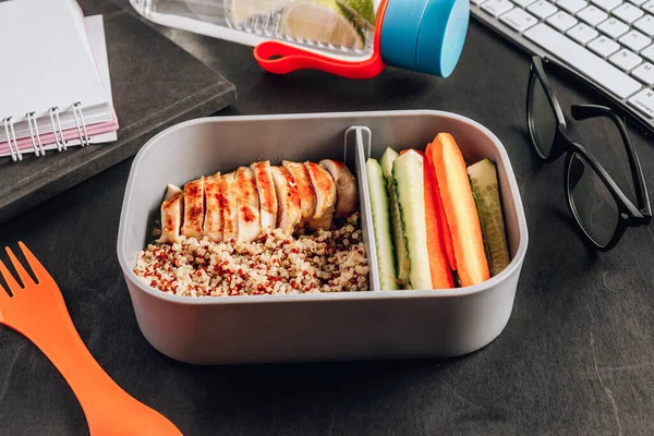 Healthy meal prep containers with quinoa, chicken and and sticks of cucumber and carrots on black wooden background. Healthy lunch at office. Take away concept. Selective focus