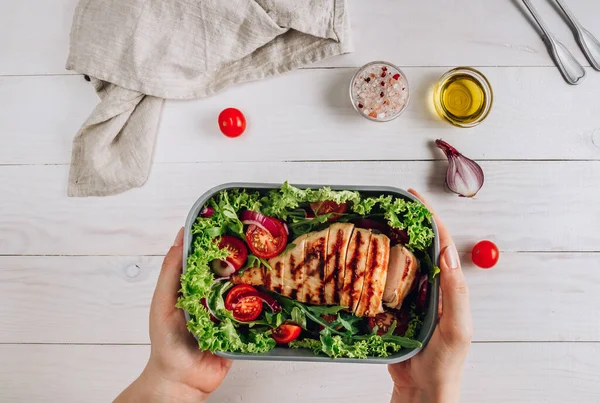 Lunch box with chicken and salad, ready to go for work or school. Meal preparation or dieting concept. White wooden table. Top view. Food container in female hands