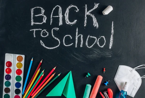 Protective mask and sanitizer, School supplies on blackboard background. Chalk inscription - Back to school. Top view. Flat lay