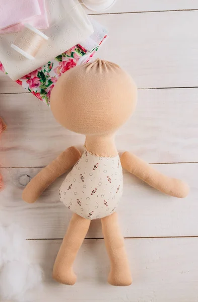 Textile doll in the sewing process with accessories and materials: fabric, thread, needle on white wooden background