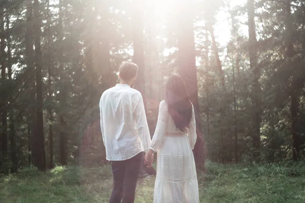 Young beautiful Slavic couple in love walks through the woods, sweet girl in a simple boho-style wedding dress. They are happy and calm. Golden hour, summer day, warm. They kiss, hug and hold hands.