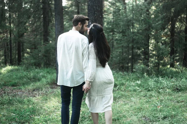 Young beautiful Slavic couple in love walks through the woods, sweet girl in a simple boho-style wedding dress. They are happy and calm. Golden hour, summer day, warm. They kiss, hug and hold hands.