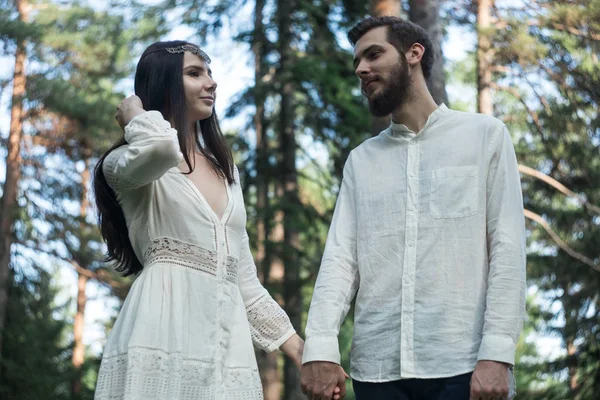 Young beautiful Slavic couple in love walks through the woods, sweet girl in a simple boho-style wedding dress. They are happy and calm. Golden hour, summer day, warm. They kiss, hug and hold hands.