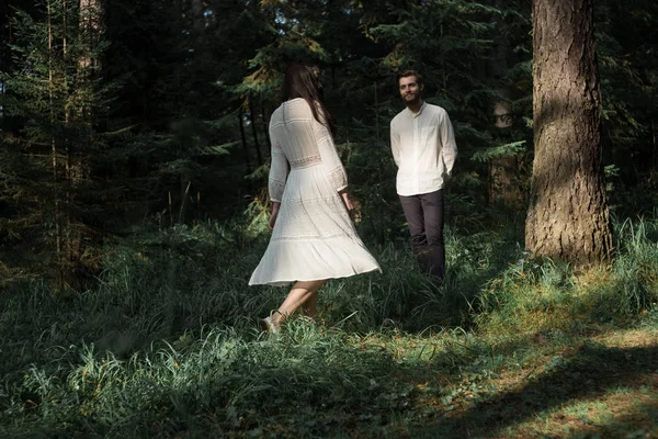 Young beautiful Slavic couple in love walks through the woods, sweet girl in a simple boho-style wedding dress. They are happy and calm. Golden hour, summer day, warm. They kiss, hug and hold hands.