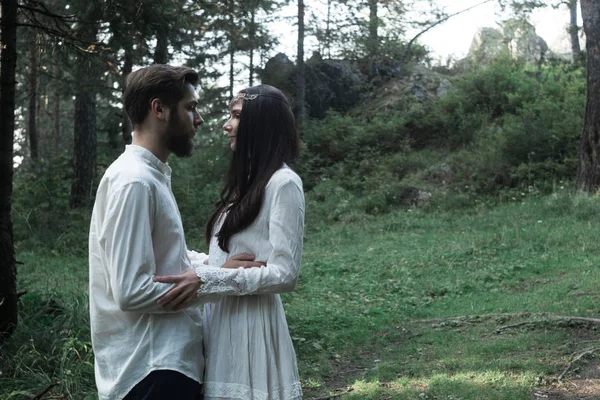 Young beautiful Slavic couple in love walks through the woods, sweet girl in a simple boho-style wedding dress. They are happy and calm. Golden hour, summer day, warm. They kiss, hug and hold hands.