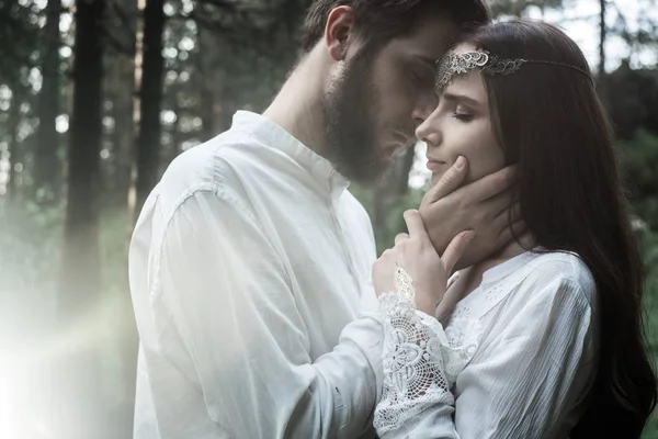 Young beautiful Slavic couple in love walks through the woods, sweet girl in a simple boho-style wedding dress. They are happy and calm. Golden hour, summer day, warm. They kiss, hug and hold hands.