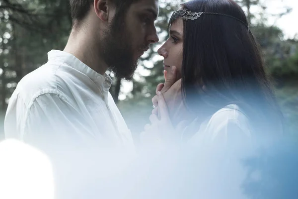 Young beautiful Slavic couple in love walks through the woods, sweet girl in a simple boho-style wedding dress. They are happy and calm. Golden hour, summer day, warm. They kiss, hug and hold hands.