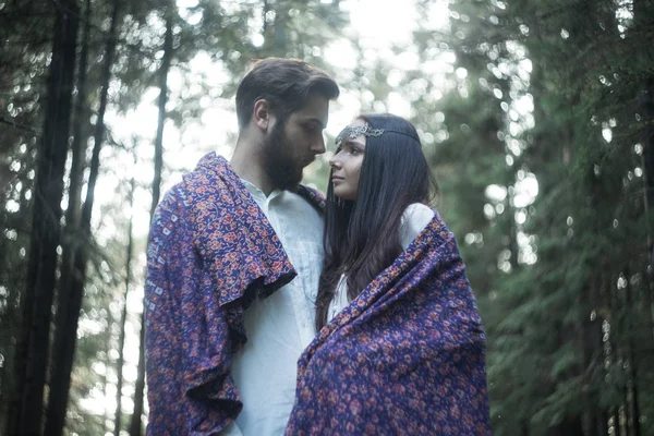 Young beautiful Slavic couple in love walks through the woods, sweet girl in a simple boho-style wedding dress. They are happy and calm. Golden hour, summer day, warm. They kiss, hug and hold hands.