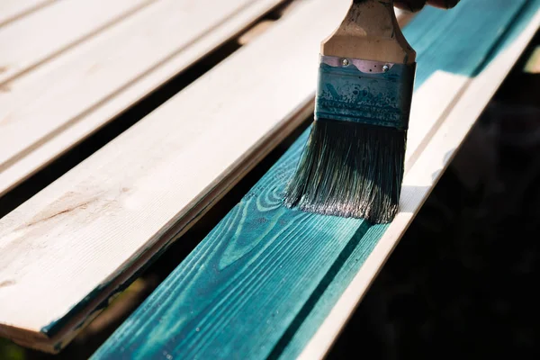 Coating with colored protective paint on the wooden surface of the lining boards. Hand with a brush paints a bright turquoise surface outdoors in Sunny weather.