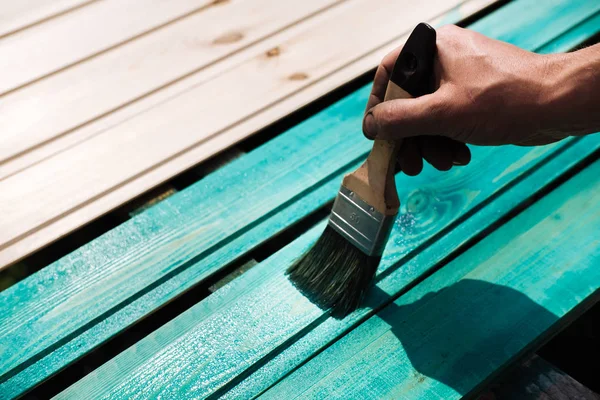 Coating with colored protective paint on the wooden surface of the lining boards. Hand with a brush paints a bright turquoise surface outdoors in Sunny weather.