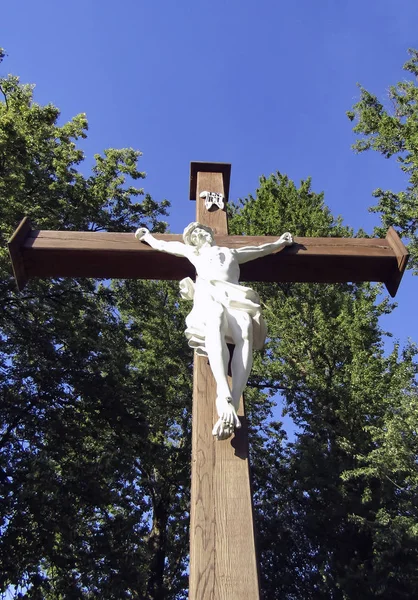 Statue crucifixion of Jesus Christ on a wooden cross against the background of blue clear sky and trees. A beautiful white sculpture depicting Jesus Christ on the christian cross.