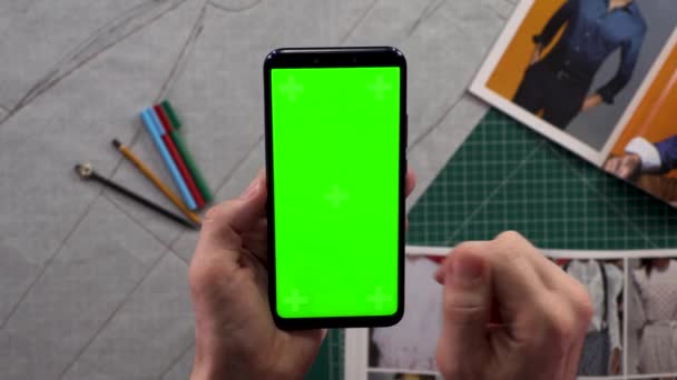 Close-up de mãos dos homens segurando tela verde vertical do telefone inteligente no guarda-roupa de fundo com a preparação de roupas. Designer criando um padrão de roupas. Designer de moda estudando desenhos . — Vídeo de Stock