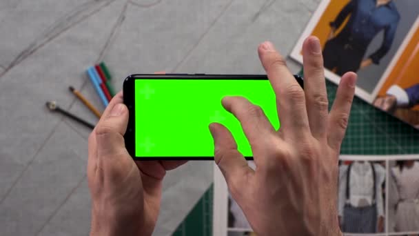 Närbild av mens händer innehav smart telefon med horisontella grön skärm i bakgrunden garderob med förbereda kläder. Modedesigner studera ritningar. — Stockvideo
