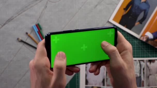 Nahaufnahme von Herrenhänden, die ein Smartphone mit einem horizontalen grünen Bildschirm im Hintergrundschrank mit vorbereiteter Kleidung halten. Bekleidungsdesigner. Modedesignerin studiert Zeichnungen. — Stockvideo