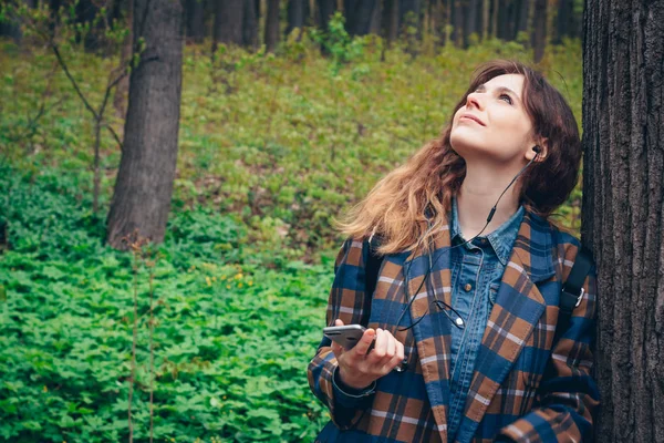 Young woman listening music at forest. Cheerful young woman walking alone in forest and enjoying music using smart phone.