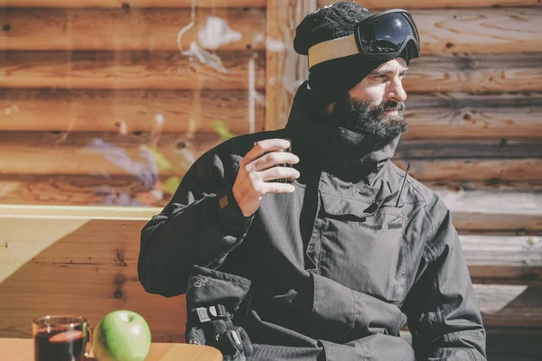 Bearded cool snowboarded taking rest after ride session.Young man drinking cup of hot tea on sunny terrace. Blurred background.Horizontal.Visual effects.