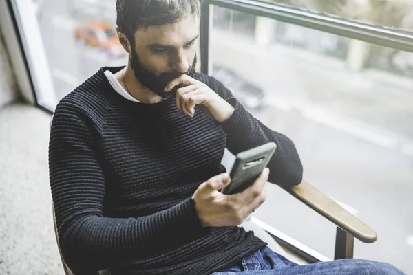 Bonito homem barbudo usando smartphone enquanto descansa no loft estúdio moderno. Fundo desfocado . — Fotografia de Stock