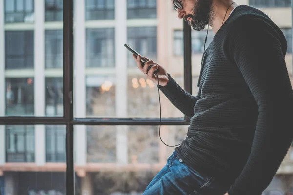 Junger bärtiger Mann, der steht und sein Mobiltelefon zum Hören digitaler Hörbücher benutzt. horizontal.panoramische Fenster auf verschwommenem Hintergrund. — Stockfoto