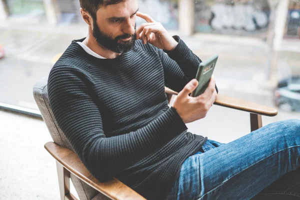 Homem barbudo pensivo sentado em casa e usando smartphone móvel. Homem usando gadget na entrada do hotel. Horizontal. Fundo desfocado . — Fotografia de Stock