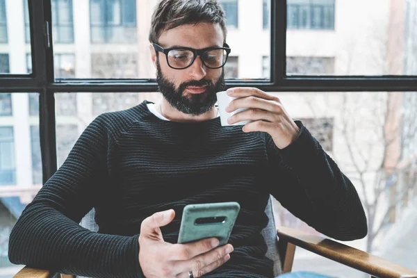 Handsome skäggig man wering glasögon dricka kaffe hemma och använder mobiltelefon. Casual avkopplande och resten tid koncept. Suddig bakgrund. — Stockfoto