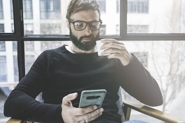 Красивый бородатый мужчина в очках пьет кофе дома и пользуется мобильным телефоном. Свободное время для отдыха и расслабления. Размытый бэкграунд. . — стоковое фото