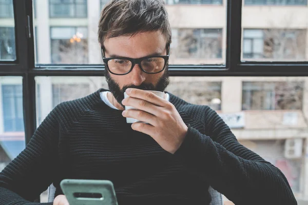 Bonito barbudo vestindo óculos e bebendo café enquanto descansa em casa. Homem usando smartphone móvel para navegar na internet. Horizontal. Fundo desfocado . — Fotografia de Stock