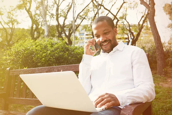 Sorrindo americano Africano empresário em roupas informais trabalhando na rua ensolarada no laptop, verificando e-mail. Homem usando smartphone para chamar amigos através de redes sociais . — Fotografia de Stock