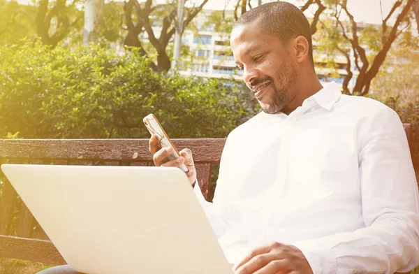 Sorrindo americano Africano empresário em roupas informais trabalhando na rua ensolarada no laptop e smartphone para chamar amigos através de redes sociais . — Fotografia de Stock