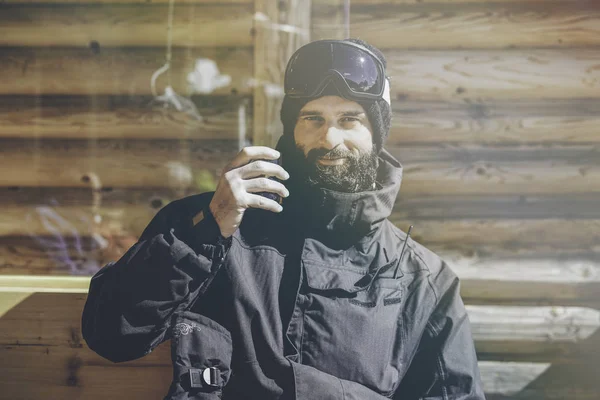 Bearded smiling snowboarded taking rest after ride session.Young man drinking cup of hot tea on sunny terrace. Blurred background.Horizontal.Visual effects. Stock Photo