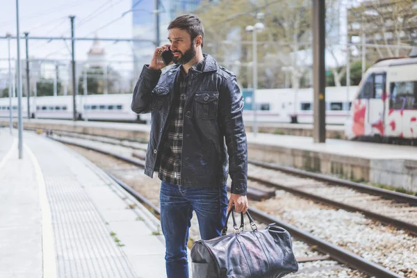 Atractivo hombre barbudo usando un teléfono inteligente en su mano está de pie con la bolsa de viaje en la plataforma esperando el tren — Foto de Stock