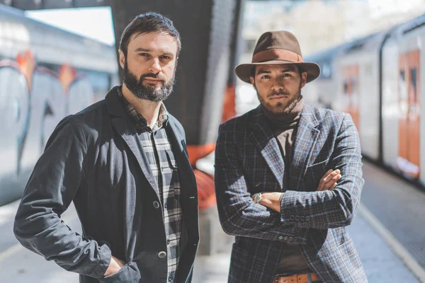 Portrait of two multi-ethnic bearded hipsters wearing together casual clothes and waiting the train on the railway platform