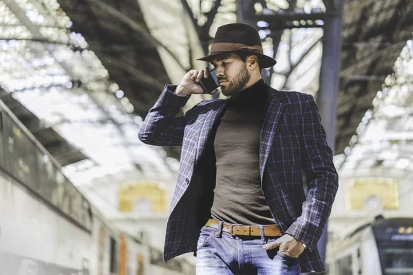 Atractivo y elegante joven hispano sosteniendo un teléfono inteligente en su mano y llamando mientras está de pie en la plataforma del ferrocarril esperando el tren — Foto de Stock