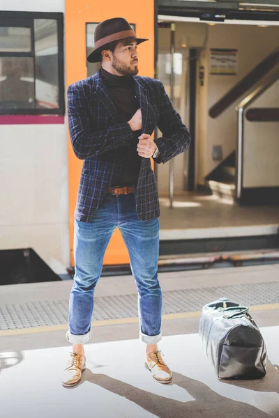Joven hombre hispano atractivo con sombrero y de pie en la plataforma del ferrocarril esperando el tren. Vertical . — Foto de Stock