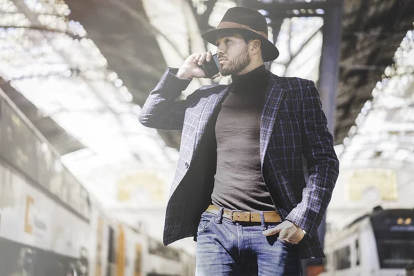 Atractivo y elegante joven hispano sosteniendo un teléfono inteligente en su mano y llamando mientras está parado en la plataforma del ferrocarril esperando el tren — Foto de Stock