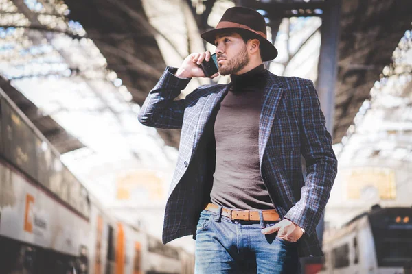 Guapo joven hispano elegante sosteniendo el teléfono inteligente en su mano y llamando mientras está parado en la plataforma del ferrocarril esperando el tren — Foto de Stock